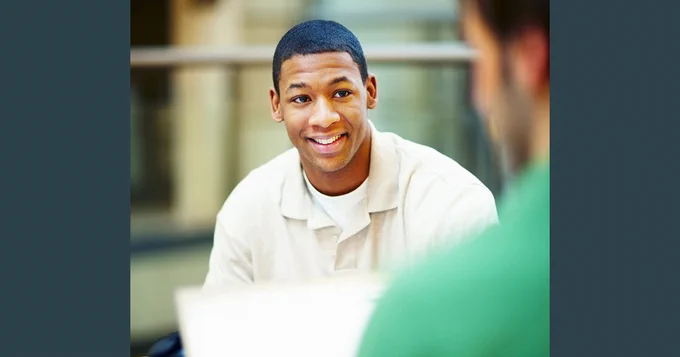 A person smiling while participating in a conversation, seated across from another person with a laptop on the table.