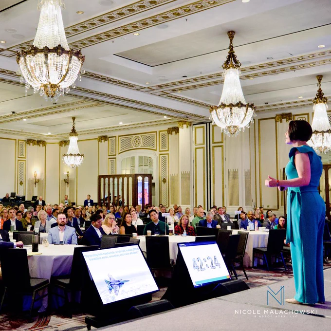Nicole presenting to a large audience in an elegant ballroom during a professional event.