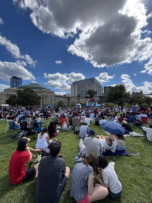 Crowd sits on U Of T front campus lawn watching soccer match on large screen.