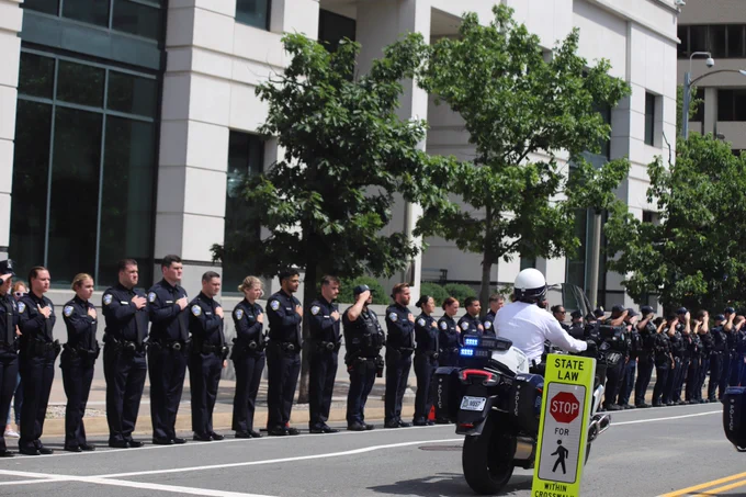 Motor officers driving down the street on motorcycles while ACPD officers stand in a line and salute.