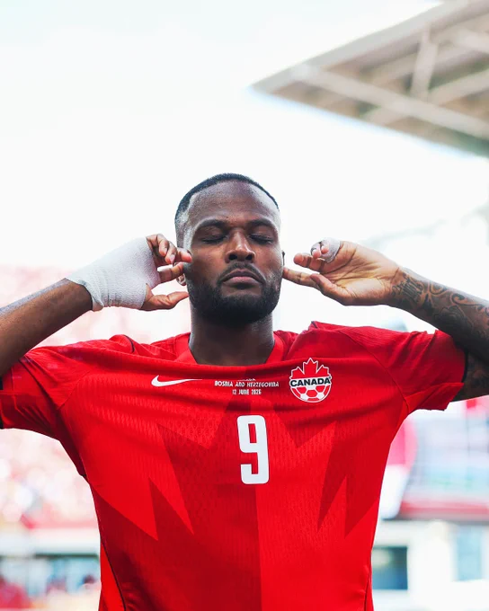 Cyle Larin celebrates scoring for Canada against Bosnia in the World Cup, with his eyes closed and both fingers on his ears