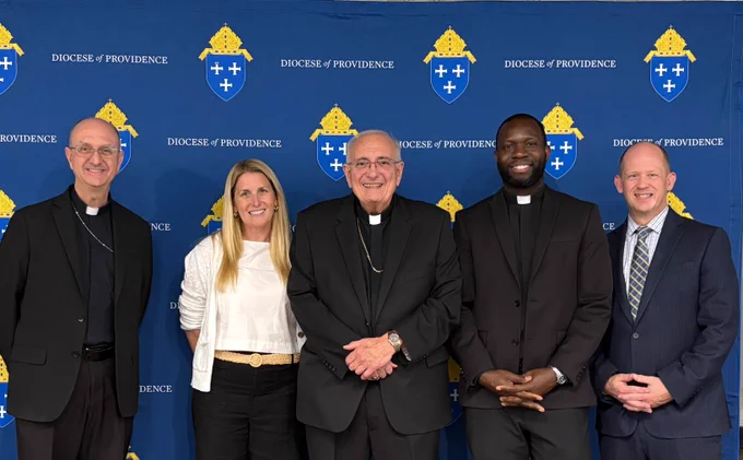 Five individuals stand in front of a "Diocese of Providence" backdrop. They are smiling for the photo and are dressed in formal and semi-formal attire.