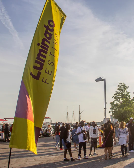 A crowd gathers at an outdoor waterfront event near a stage, with a tall yellow flag reading “Luminato Festival” in the foreground. The Protect Ontario logo appears in the corner.