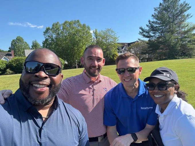 Four friends are taking a selfie on a sunny day at a golf course. They are smiling and dressed in casual or sporty attire, enjoying the outdoors.