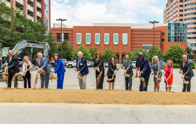 Group of people participating in a groundbreaking ceremony, each holding shovels. The scene is outdoors with a construction vehicle and buildings in the background.