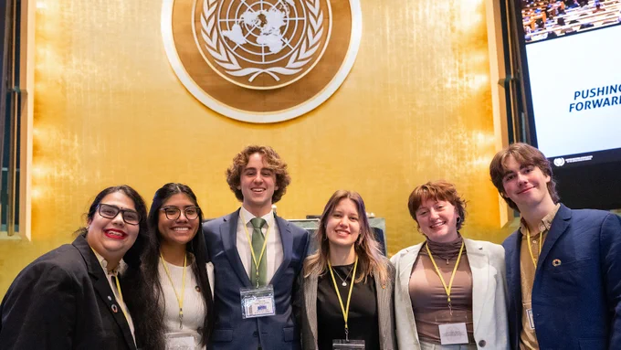A group of six people stands smiling in front of a large emblem of the United Nations. They are inside a formal setting, and most are wearing business attire with lanyards.