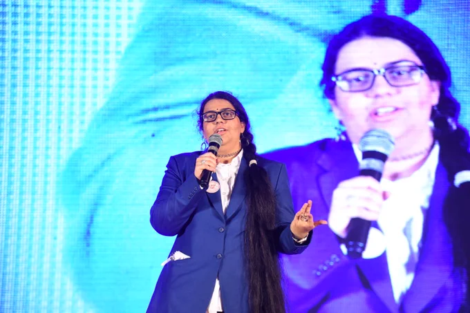 A person in a blue suit speaks on stage while holding a microphone. A large screen behind them shows their image.