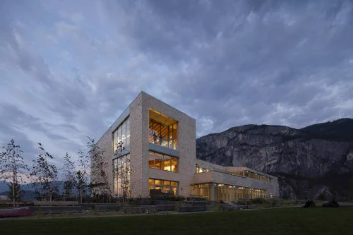 A modern building with large glass windows glows warmly at dusk, set against a backdrop of rugged mountains and a cloudy sky.