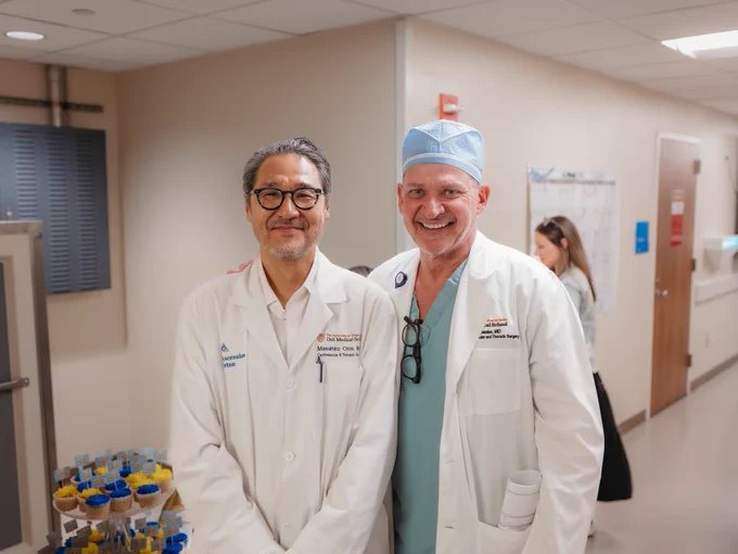 Two surgeons pose for a photo in a hospital setting. They are both wearing white coats.