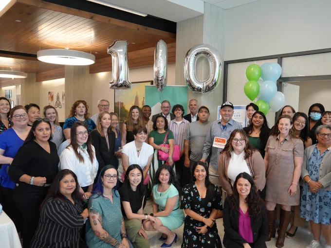 A large group of people pose for a photo at a celebratory event. Balloons that read "100" are visible.