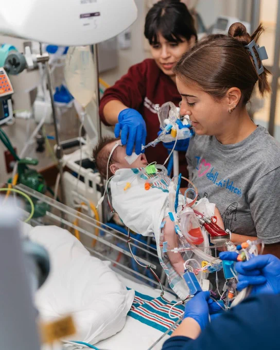 Nurses handle an infant connected to medical devices in a hospital setting.