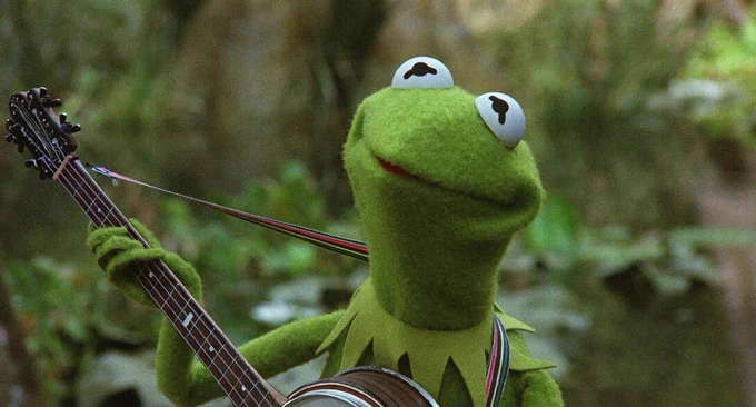A close-up of the Kermit the Frog puppet, holding a banjo in a swamp. (Courtesy)