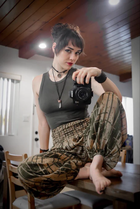 Hailie is sitting on their table, taking a mirror selfie with their camera. They look into the lens while wearing a green tank top and big loose pants.