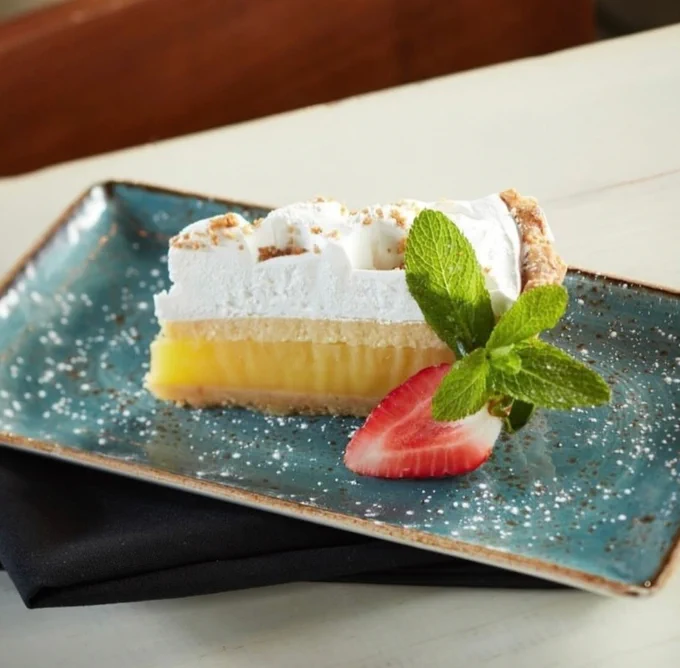 Slice of lemon meringue pie topped with toasted meringue, garnished with a fresh strawberry half and mint leaves on a blue ceramic plate.