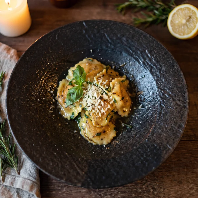 Black plate with four ravioli pieces topped with grated cheese and fresh basil on a wooden table.