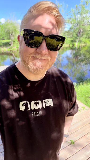 Selfie of me in the backyard of a cottage where a river and trees are in the background. I’m wearing a pair of sunglasses and a black shirt that has faces of Batman from Batman the animated series on it.