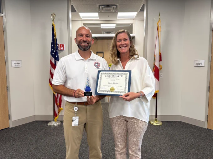 Two individuals standing in an office hallway holding a certificate and an award, with American and state flags behind them.