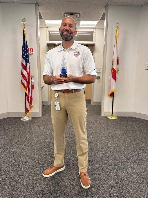 Person in khaki pants and white polo holding a blue glass award standing between American and Florida state flags indoors.