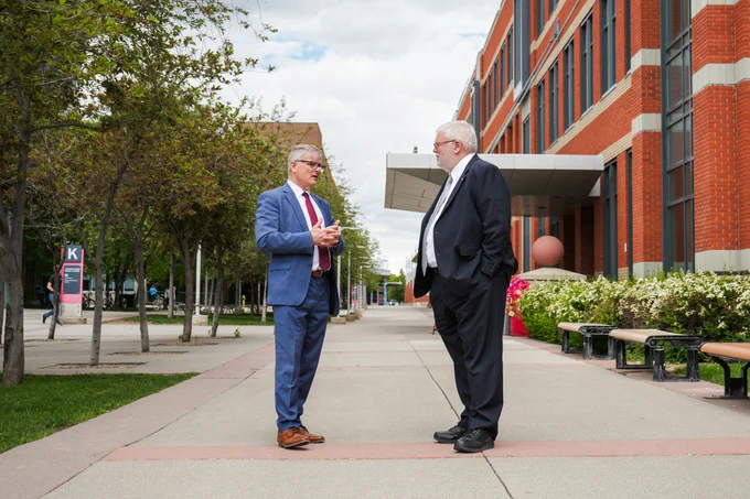 Two individuals in business attire are conversing outside a brick building on SAIT campus. The walkway is lined with trees and benches, and there's a sign in the background.