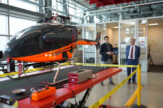 A small helicopter drone with a black and orange design is displayed indoors on a table, surrounded by caution tape. Two people are standing nearby, observing the drone. The room has large windows and office cubicles in the background.