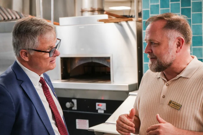 Two individuals are having a conversation in an industrial kitchen setting. One is dressed in a suit and tie, and the other is wearing a polo shirt. An industrial oven in the background and teal tiles are visible.