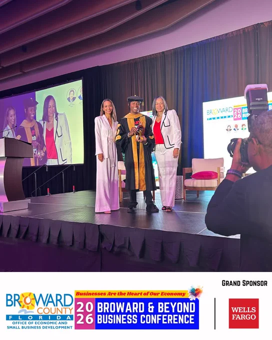 Three smiling people standing on a stage with the person in the middle wearing robe and hat and holding an award being photographed by a photographer standing in front of the stage.