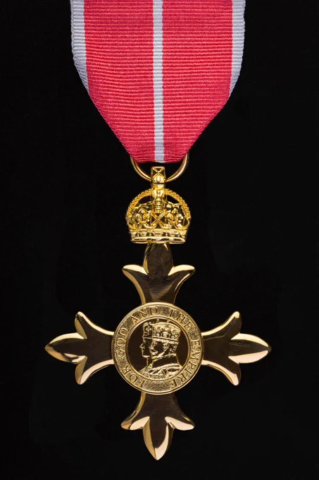 A medal with a red and white striped ribbon is hanging against a black background. The medal is cross-shaped and features the profile of a monarch surrounded by the words "For God and the Empire." A crown sits above the cross, connecting it to the ribbon.
