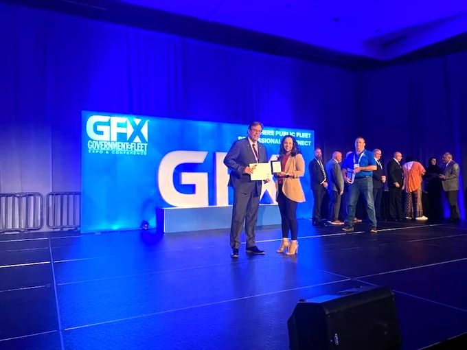Two people pose for a photo on a stage holding plaques. There are people lingering behind them.