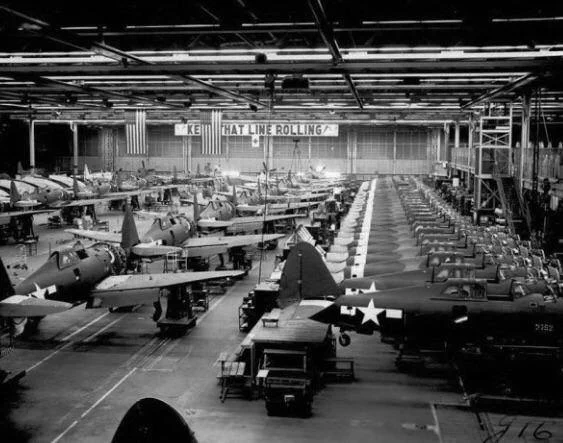 A large aircraft manufacturing facility with rows of military planes in progress. An overhead sign reads “KEEP THAT LINE ROLLING.” American flags are displayed in the background.