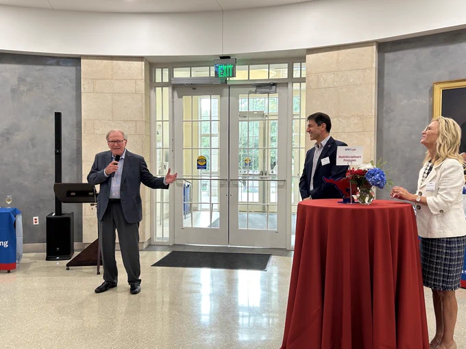 Dr. Bobby B. Lyle stands in front of the podium at an SMU Lyle School of Engineering admitted students event giving remarks with his hands outstretched and holding a microphone. Dean Jalili looks on at him from the side and smiles. 