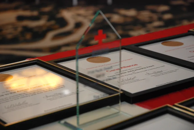 A table displays framed certificates and an acrylic award in the shape of a house. A red cross symbol is visible in the background.