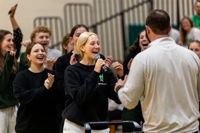 A lively group of people in casual attire celebrating inside a gymnasium. A woman at the center holds a microphone, engaging with a man facing away.