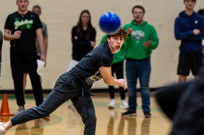 A young man in a gym launching a blue dodgeball energetically. Onlookers in the background watch intently, adding a competitive atmosphere.