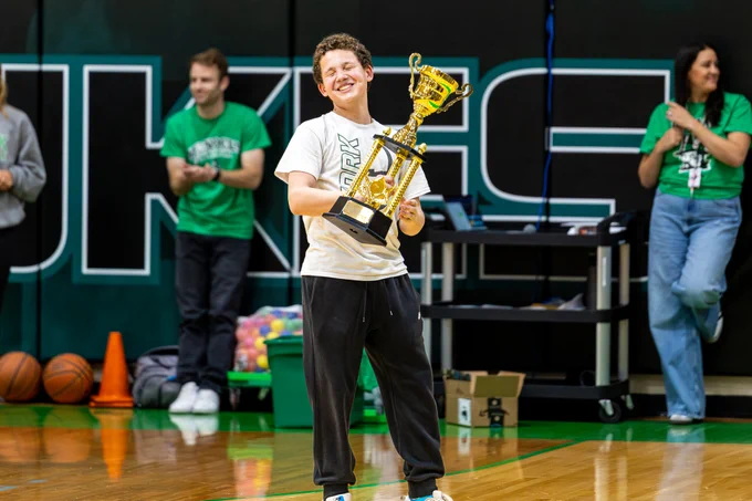 A smiling young individual holds a large gold trophy in a gymnasium, indicating a joyous achievement. People in the background applaud and smile, contributing to the celebratory atmosphere.