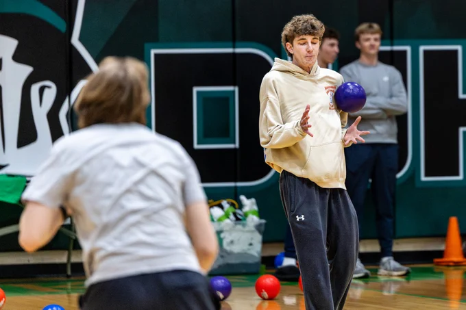 A young man in a hoodie braces for impact as he plays dodgeball in a gym. The atmosphere is tense and lively.