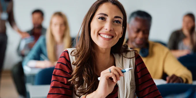 Student in a striped shirt attentively holding a pen during a class with peers seated behind them.