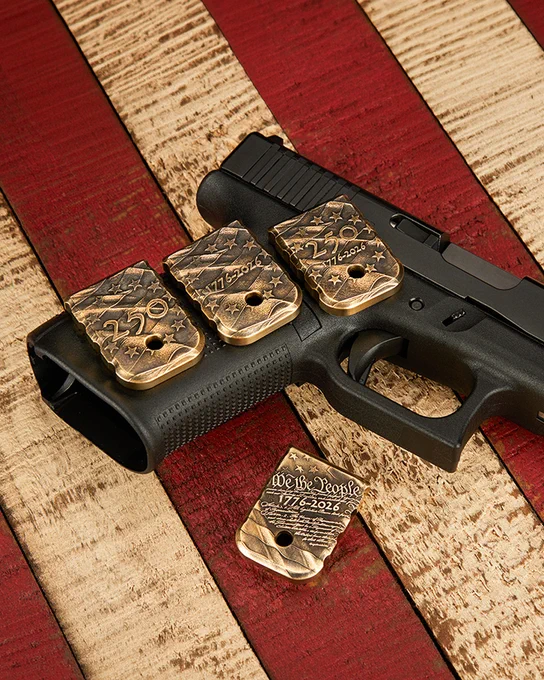 Four engraved brass magazine base plates commemorating America's 250th anniversary displayed on and beside a black pistol. The designs feature patriotic stars, waving flag elements, "250," "1776-2026," and "We the People" engravings against a distressed American flag wood background.