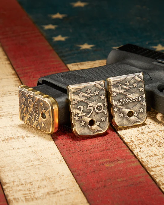 Three engraved brass magazine base plates shown installed on pistol magazines, highlighting the patriotic 250th anniversary designs. The plates feature raised stars, flag-inspired textures, and commemorative engravings against a weathered American flag backdrop.
