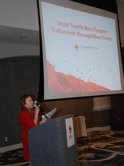 A person stands at a podium with a microphone in a conference room. Behind them, a large screen displays "2026 North Bay Chapter Volunteer Recognition Event" with the American Red Cross logo.