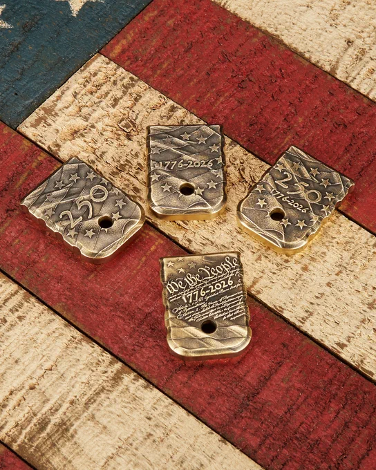 Four antiqued brass magazine base plates arranged on a rustic American flag wood surface. Each plate features detailed patriotic engravings including stars, waving flag patterns, "250," "1776-2026," and a Constitution-inspired "We the People" design commemorating America's 250th anniversary.