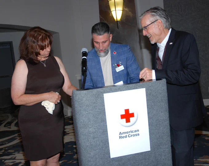 Three individuals stand behind a podium with an American Red Cross logo. They appear to be in conversation, with one person holding a paper and another holding a cloth. The setting appears to be an indoor event space.