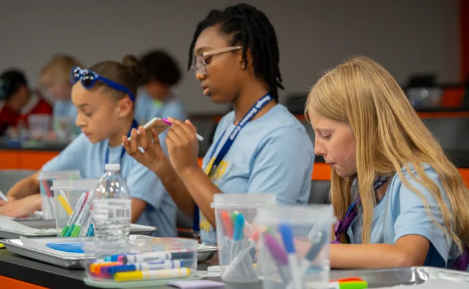 Three girls sitting a lecture hall desk and coloring their own tiles as part of the art portion of the camp. They all wear light blue shirts, but the girl closest to the camera has long, blonde hair, unlike the other girls who both have short, dark hair. They each have a small tub of markers in front of them.
