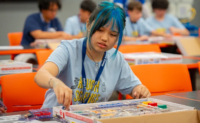 A girl with blue dyed bangs works on a similar board. She sits by herself in the frame with a group of four boys two rows behind her.