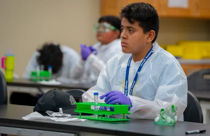 A young man in a disposable lab coat sitting at a desk, listening to the instructor speak out of frame. he wears a blue lanyard, purple latex gloves, and has biology lab equipment in front of him. There are two other boys in the background, dressed the same.