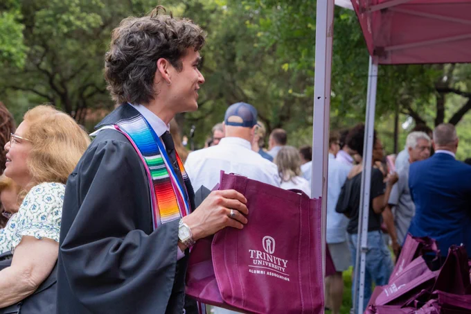 A graduate holds a maroon Trinity University swag bag outside.
