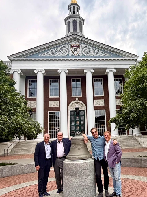 Four people stand in front of a large building with columns and a bell. The building has an emblem at the top and resembles the architecture of Harvard Business School. Trees flank the building on both sides.