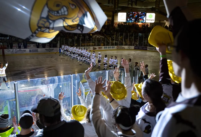 A hockey game setting with players from the Western Michigan team lined up on the ice. The audience, with some wearing yellow hats, is cheering energetically. A large Western Michigan flag is visible in the foreground. The arena is well-lit, and a scoreboard with advertisements is visible above.