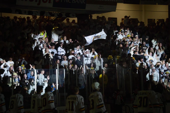 A group of hockey spectators known as the Lawson Lunatics fills a section of seats, many wearing white clothing. One person is waving a flag. Below, a few hockey players in uniform face the crowd. Advertisements for Meijer and PNC Bank are visible above the seats.