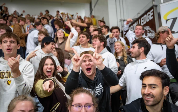 A large group of enthusiastic WMU fans are cheering in Lawson Arena with many wearing team apparel. People are raising their hands and pointing, showing excitement. 