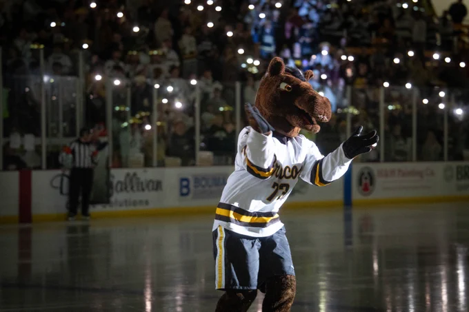 Buster the Bronco is performing on an ice rink, wearing a jersey, surrounded by a crowd with illuminated phones. The setting appears to be at Lawson Arena, where people are watching the mascot with excitement.